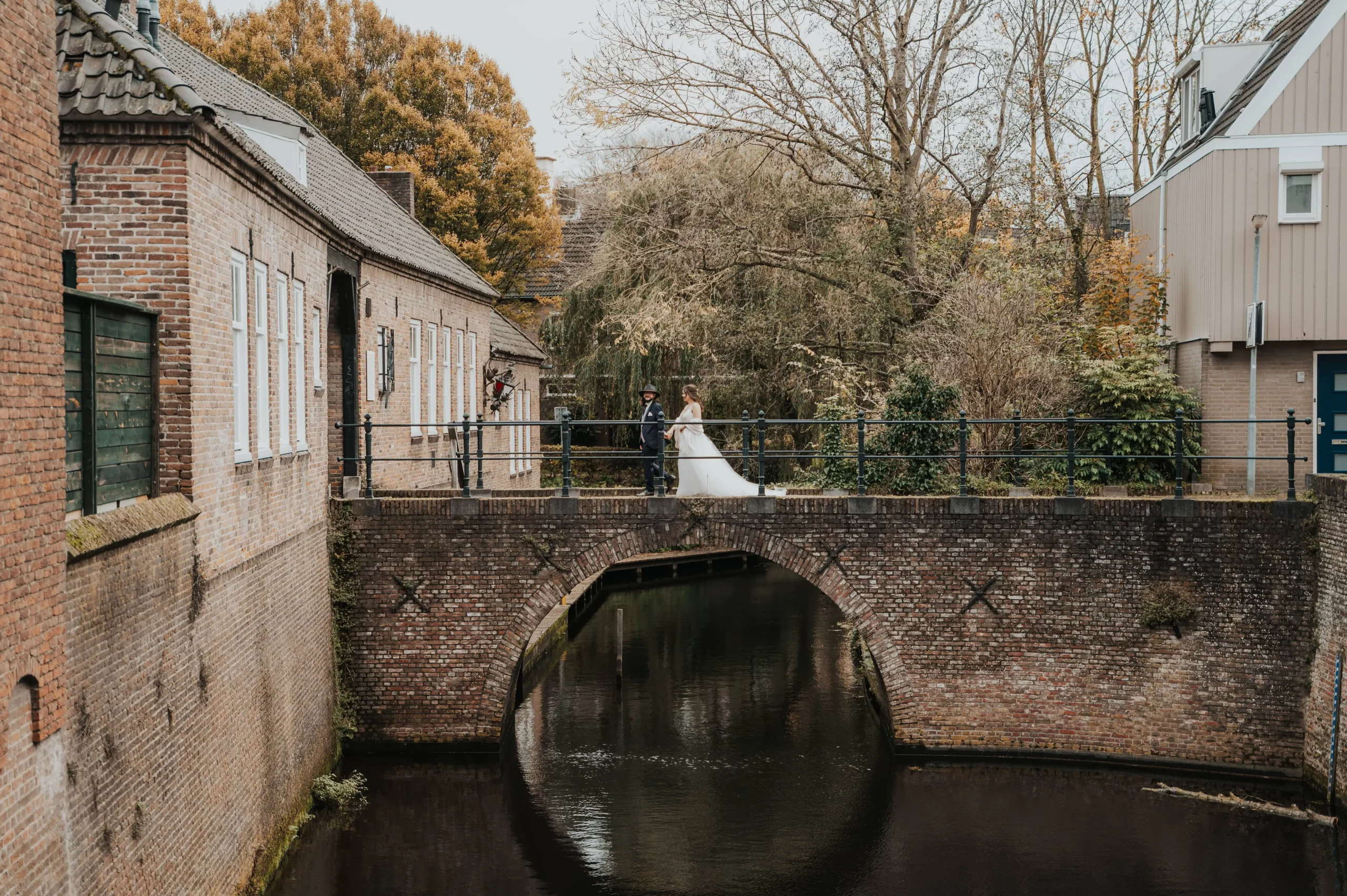 Bruidspaar wandelt over een stenen brug boven een waterloop, tussen huizen en bomen