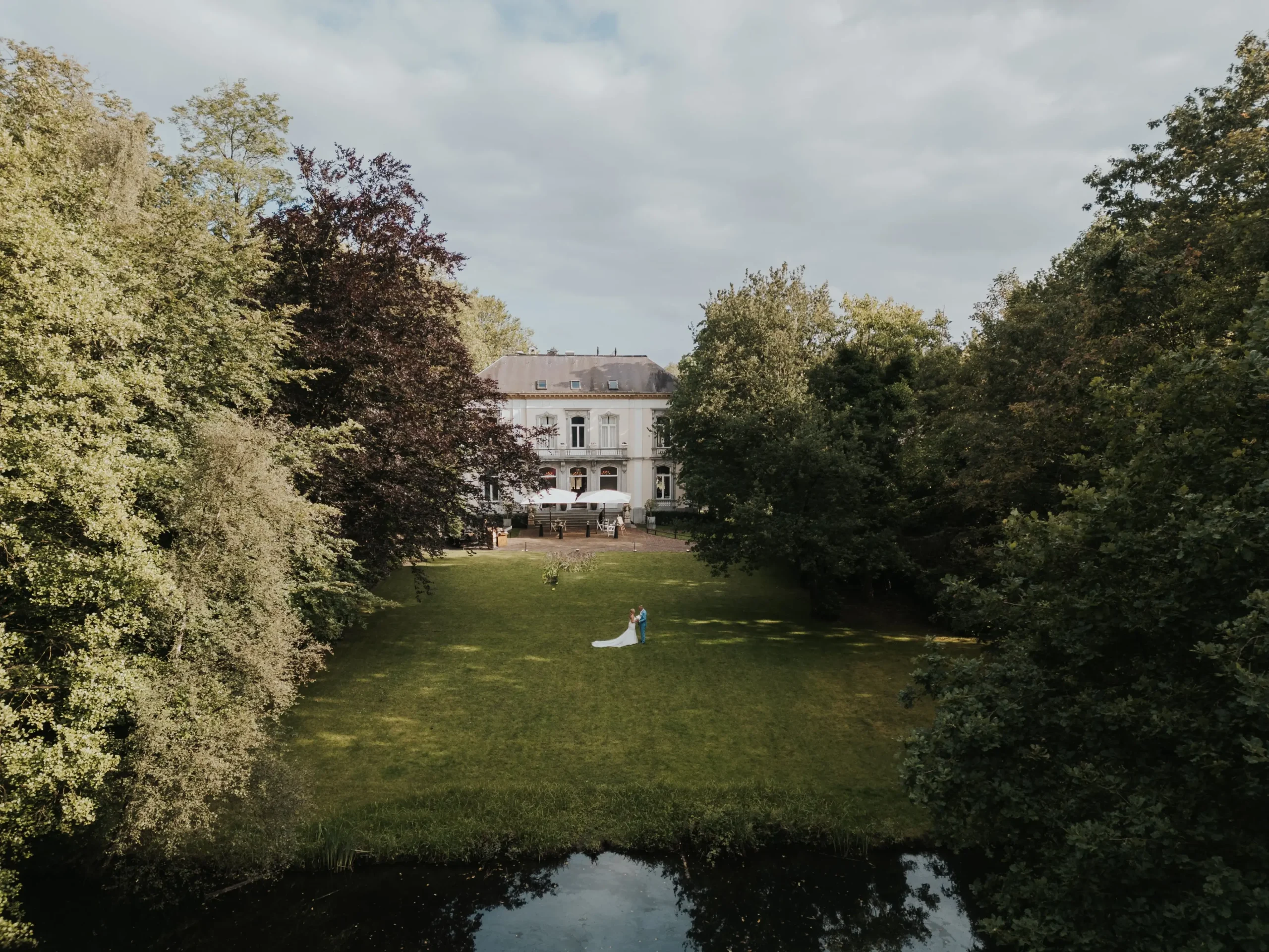 Bruidspaar klein in beeld op een gazon bij een lichtgekleurd landhuis, bomen en water op de voorgrond.